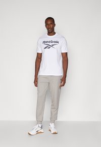 Bijela majica kratkih rukava s tamnoplave Reebok oznake, uparena s svijetlosivim tapered trenirkama i bijelim tenisicama s naglascima.
