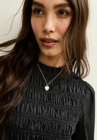 Czarna bluzka z marszczeniem i bufkami, z naszyjnikiem w kształcie srebrnego serca. Gładkie włosy o naturalnym układzie, na jasnym tle.