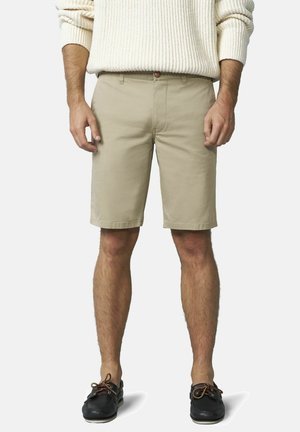 Man die beige knielange korte broek, een crèmekleurige trui en zwarte bootschoenen draagt, staand met ontspannen handen langs het lichaam.