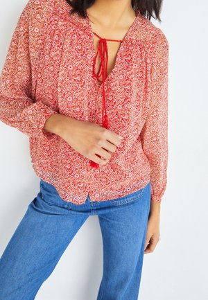 Femme portant un chemisier rouge à fleurs à manches longues avec un nœud devant et un jean bleu, debout sur un fond blanc.