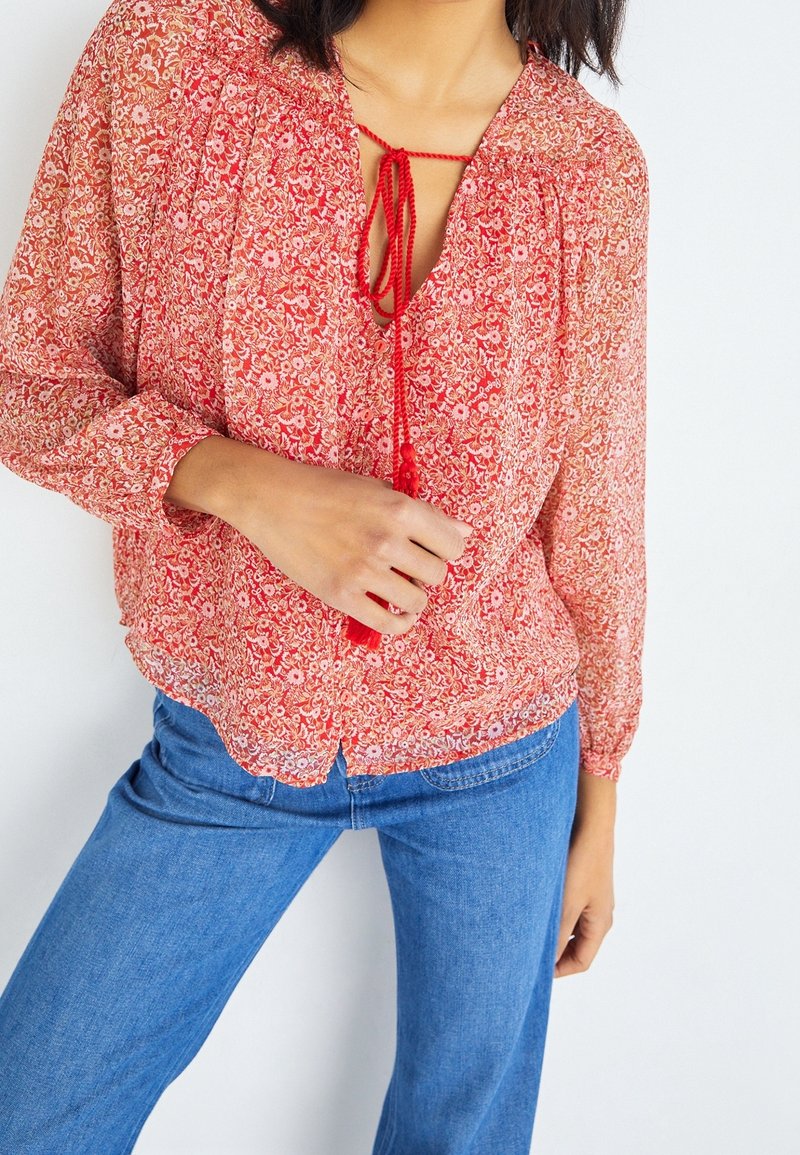 Femme portant un chemisier rouge à fleurs à manches longues avec un nœud devant et un jean bleu, debout sur un fond blanc.