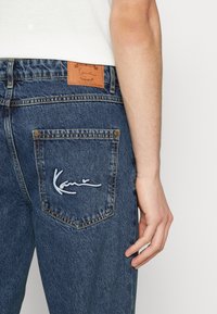 Jeans de mezclilla azul oscuro con un parche de cuero en la cintura y detalle de firma bordado en el bolsillo trasero. Diseño clásico de cinco bolsillos.
