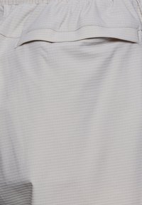 Hellgrauer Stoff mit kleinem Rautenmuster, das eine horizontale Klappentasche nahe dem oberen Bereich und einen elastischen Bund darüber aufweist.