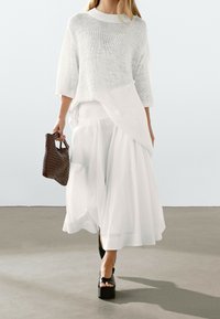 Witte gebreide trui met 3/4 mouwen, gelaagd over een witte flared maxi rok. Zwarte platform sandalen en een gevlochten bruine handtas maken de look compleet.