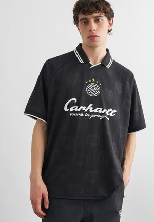 Giovane uomo che indossa una polo Carhartt nera con bordi e loghi bianchi, in piedi davanti a uno sfondo chiaro uniforme.