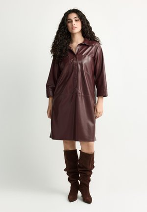 Woman with curly hair wearing a knee-length dark brown leather shirt dress and matching slouchy suede boots standing against a plain background.