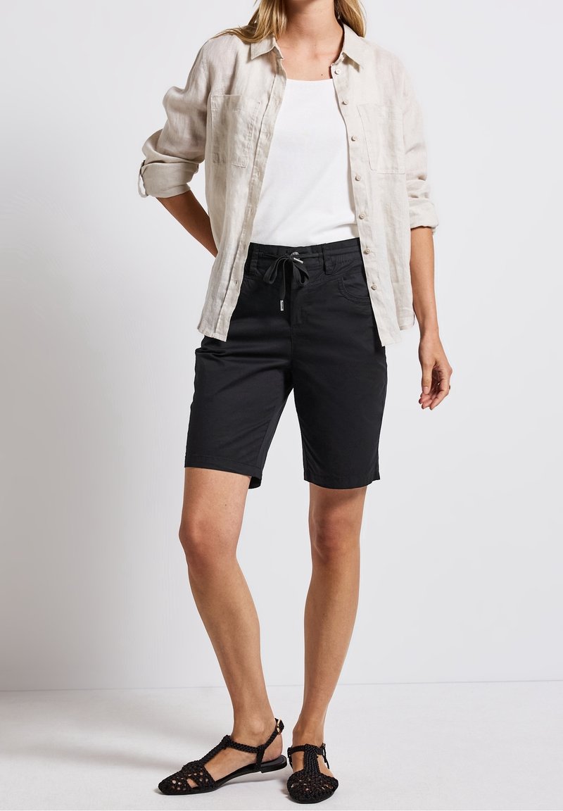 Femme portant une chemise beige à boutons sur un haut blanc, un short noir à cordon et des sandales tissées noires, debout devant un fond uni.