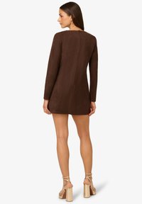 Hnědé šaty z umělé suroviny s dlouhými rukávy, jednoduchého střihu, se zipem na zadní straně a délkou nad kolena, doplněné béžovými sandály na vysokém podpatku.