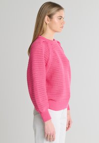 Różowy sweter z dzianiny z teksturowanym wzorem, okrągłym dekoltem i ściągaczami na rękawach. Modelka nosi jasne spodnie, pokazana w profilu.