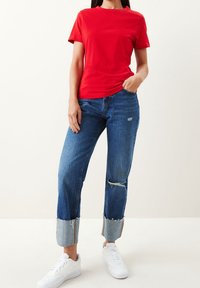 Person i en rød kortærmet skjorte, opfoldede slidte blå jeans med huller på knæene og hvide sneakers, står med den ene hånd i lommen.
