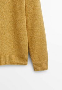 Musztardowożółty dziergany sweter o fakturowanym wykończeniu, z prążkowanymi mankietami i krawędzią. Tkanina wydaje się miękka i ciepła.