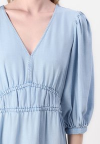 Rochie din denim de culoare bleu deschis, cu decolteu în V, mâneci bufante și talie elastică cu material plisat. Textură netedă cu detalii subtile de cusătură.