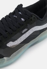 Sapatilha de malha preta com uma sola de borracha translúcida, apresentando uma faixa branca da Vans e atacadores pretos. O design inclui pormenores em tecido e uma etiqueta com o logótipo.