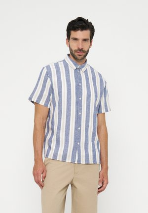 Homme portant une chemise à manches courtes boutonnée avec des rayures verticales bleues, blanches et beiges, associée à un pantalon beige, debout sur fond blanc.