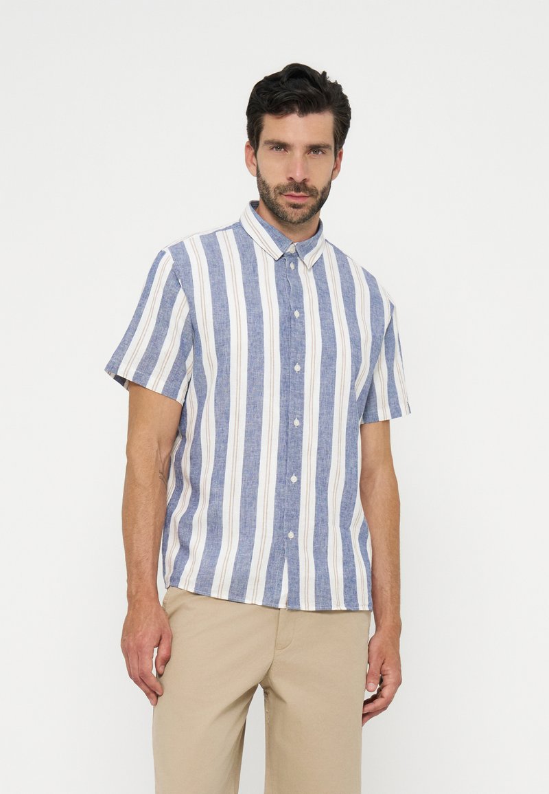 Homme portant une chemise à manches courtes boutonnée avec des rayures verticales bleues, blanches et beiges, associée à un pantalon beige, debout sur fond blanc.