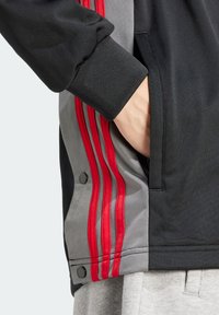 Svart idrottsjacka med grå detaljer, som har tre röda ränder längs sidan, en ficka med dragkedja och design med manschettärmar.