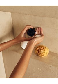 Bottiglia di vetro trasparente con pompa nera che eroga un liquido bianco, posizionata vicino a una spugna gialla naturale su una superficie beige testurizzata.