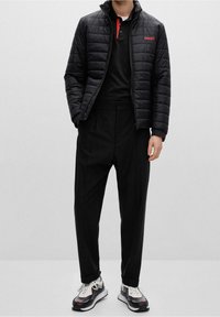 Czarna pikowana kurtka puchowa z wysokim kołnierzem i czerwonym logiem "HUGO", zestawiona z czarnymi spodniami i białymi trampkami z czerwonymi akcentami.
