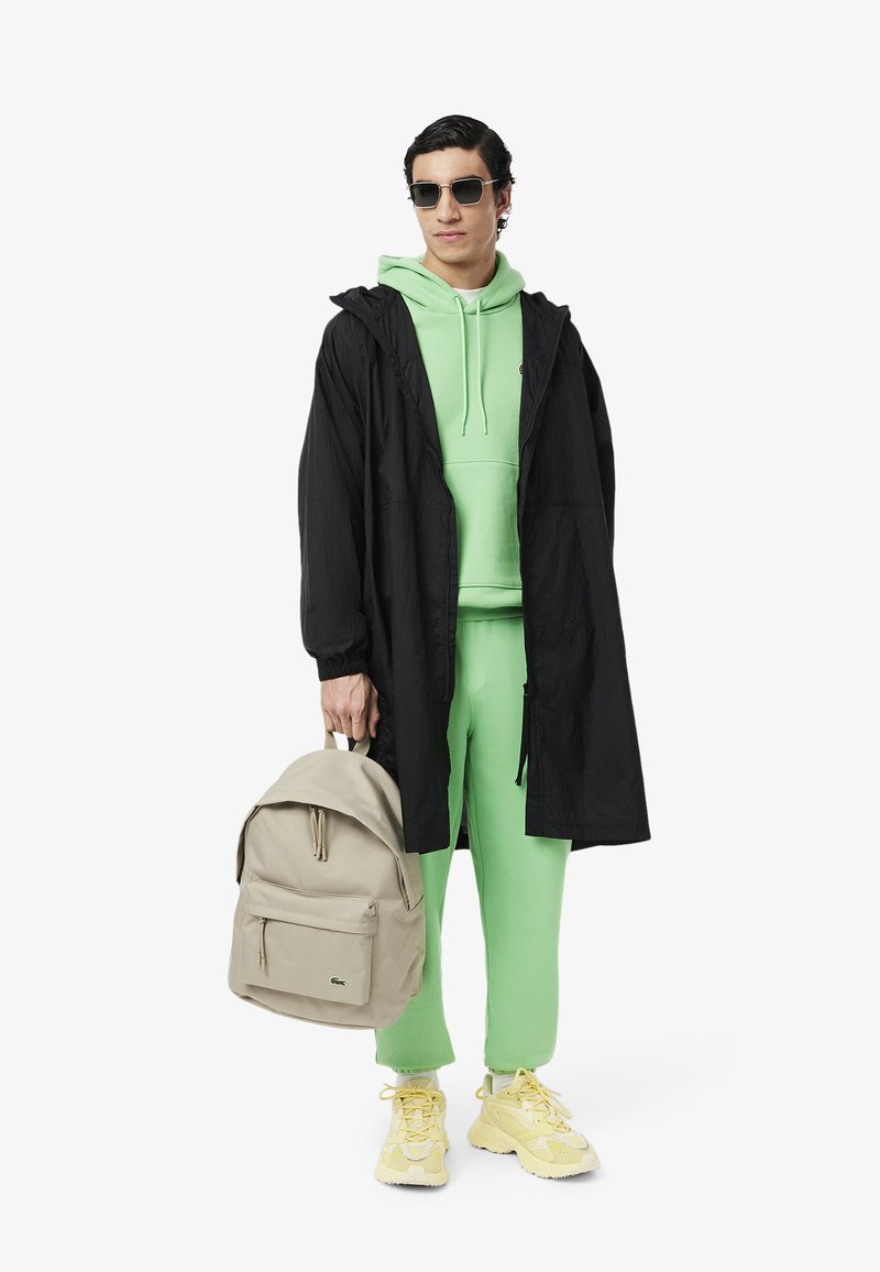 Mężczyzna w czarnej długiej kurtce na zielonym kapturze i spodniach, trzyma beżowy plecak, w żółtych sneakersach i okularach przeciwsłonecznych, stojący na białym tle.