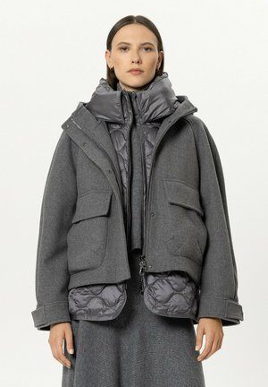 Young woman wearing layered gray quilted and wool jackets with large pockets, standing against a plain light background.