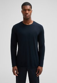 Mann in einem schlichten, dunklen Langarmshirt und dunkler Hose, steht vor einem neutralen grauen Hintergrund und schaut direkt nach vorne.