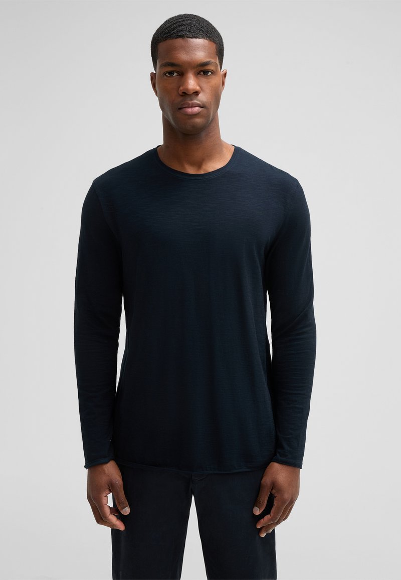 Mann in einem schlichten, dunklen Langarmshirt und dunkler Hose, steht vor einem neutralen grauen Hintergrund und schaut direkt nach vorne.