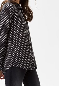 Svart skjorta med vita prickar, lång ärm, knäppning och krage. Vita prickar med jämna mellanrum; lätt tyg med en slät yta.