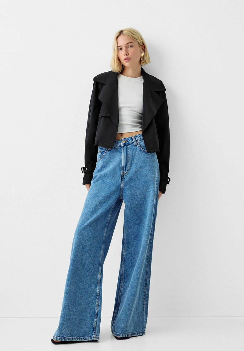 Giacca nera corta con ampi revers sopra un crop top grigio chiaro, abbinata a jeans larghi a vita alta di un blu chiaro.