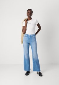 T-shirt bianca aderente con testo, jeans a campana blu chiaro e scarpe nere. La modella tiene una giacca beige sulla spalla. Sfondo semplice.