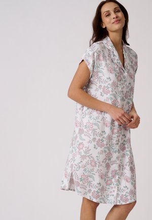 Femme portant une robe fleurie blanche longueur genou avec manches courtes et boutons, debout devant un fond clair uni.