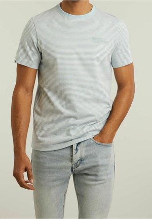Mężczyzna ubrany w jasno-niebieski, cienko prążkowany t-shirt z krótkim rękawem oraz jasnoniebieskie dżinsy, z jedną ręką w kieszeni.