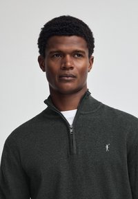 Mörkgrå zip-up tröja med hög krage och en liten broderad logotyp på bröstet. Mjuk textur och figursydd design.