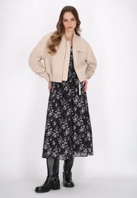 Giacca bomber beige con polsini a costine, sopra un abito maxi nero a fiori. Stivali neri al ginocchio con dettaglio in fibbia completano l'outfit.