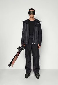 Στολή για σκι που περιλαμβάνει μαύρο ανθεκτικό στο νερό μπουφάν, μαύρο μακρυμάνικο πουκάμισο, παντελόνι χιονιού cargo και μπότες σκι, κρατώντας ένα ζευγάρι σκι.