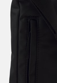 Zaino in tessuto nero con tasca con zip, design elegante e dettaglio di cucitura discreto. L'etichetta del marchio è visibile sul lato.