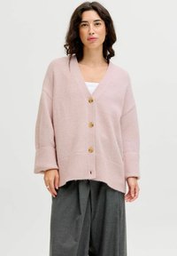 Cárdigan rosa claro con escote en V, cuatro botones de madera, puños acanalados y dobladillo. Lleva puesto sobre una camiseta blanca y pantalones de pierna ancha grises.