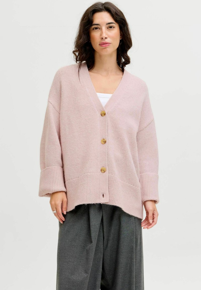 Cárdigan rosa claro con escote en V, cuatro botones de madera, puños acanalados y dobladillo. Lleva puesto sobre una camiseta blanca y pantalones de pierna ancha grises.