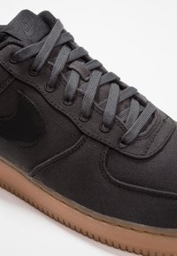 Černá plátěná teniska se šedými tkaničkami, černým logem Swoosh a podrážkou z přírodní gumy barvy tan. Má zesílené prošití a hladkou texturu.