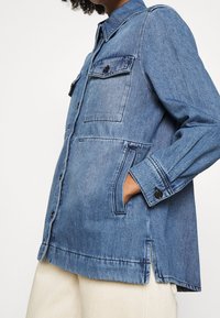 Chaqueta de denim en azul claro, con dos bolsillos en el pecho, aberturas laterales, puños con botón y cuello. Ajuste ligeramente holgado.