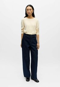 Světlá béžová košile s dlouhým rukávem v kombinaci s tmavě modrými džínami s širokými nohavicemi. Džíny mají klasické kapsy a viditelné švy na zadní straně.