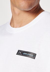 Bijela pamučna majica s crnim pravokutnim oznakom s natpisom "Under Armour" i logom, okruglim izrezom i glatkom teksturom.