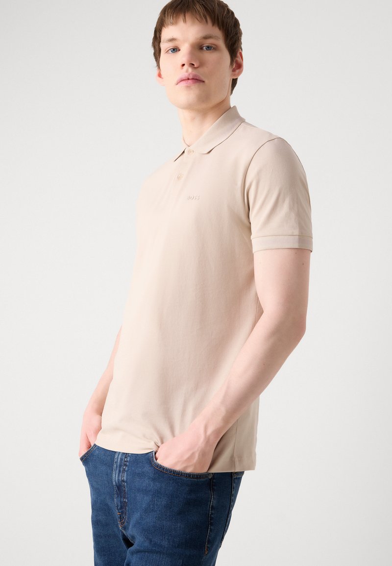 Polo beige en tissu doux, avec un col, des manches courtes et un logo sur la poitrine. Porté avec un jean bleu.