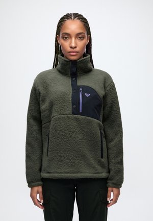 Un mannequin aux cheveux tressés porte un pull en polaire vert olive à col montant, avec un empiècement noir sur la poitrine, une fermeture éclair violette et un pantalon noir.