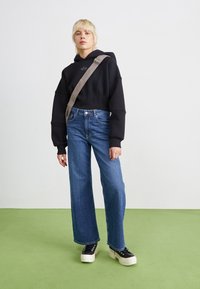Črna skrajšana kapuca, modri široki jeans hlače in črne platforme s belimi podplati. Ozadje je svetle barve z zelenim podom.