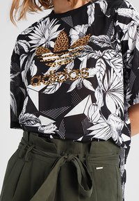 Svartvit t-shirt med blommönster och gyllene Adidas-logotyp. Kortärmad, rund halsringning, kombinerad med olivgröna byxor med bälte.