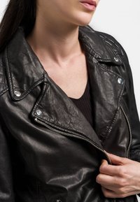 Veste de moto en cuir noir avec quincaillerie argentée, couture détaillée et fermeture éclair. Surface texturée et larges revers.