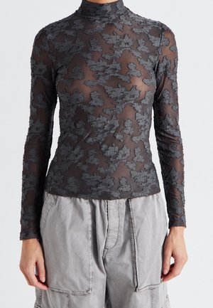 Kvinde iført sort gennemsigtig langærmet top med teksturerede blomsterrelieffer og lysegrå højtaljede cargobukser, stående mod hvid baggrund.