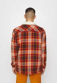 Koszula flanelowa w brązowy, czerwony i żółty wzór w kratę. Posiada miękki biały kołnierzyk i guzikowe mankiety. Regularny krój i długie rękawy.