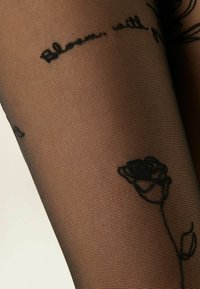 Czarne przezroczyste rajstopy z haftowanymi czarnymi różami i detalem w postaci kursywy na nodze osoby.