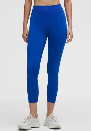 Frau trägt leuchtend blaue, hoch taillierte kurze Leggings und weiße Sportschuhe, steht vor einem einfarbigen Hintergrund.